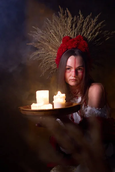 Yüzü boyalı folklor kıyafeti giymiş gizemli bir kadın koyu, dumanlı arka planda yanan bir mum tepsisi tutuyor büyüleyici ve ruhani bir atmosfer yaratıyor. .
