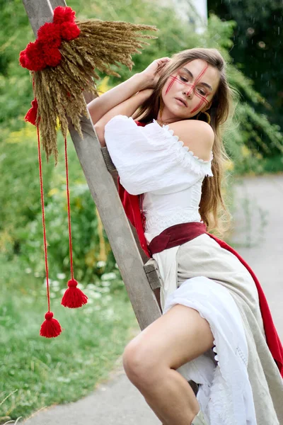 Kırmızı yüzlü bir Ukraynalı güzel, ahşap bir merdivenin üzerinde yemyeşil yaprakların ve yağmurun zeminine yaslanmış, yağmur altında yağmura karşı gelenek ve doğal güzelliğin bir karışımını sergiliyor. 