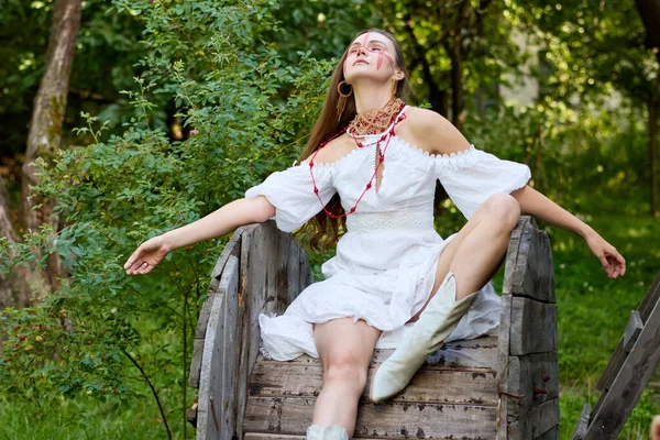 Kırmızı yüz boyası ve doğada bohem kıyafetleri olan genç bir kadın, beyaz bir elbise ve kovboy çizmeleri giyiyor, kırsal bir tahta makarada oturuyor yemyeşil bir zemin üzerinde, güneş ışığı içinden süzülüyor. .