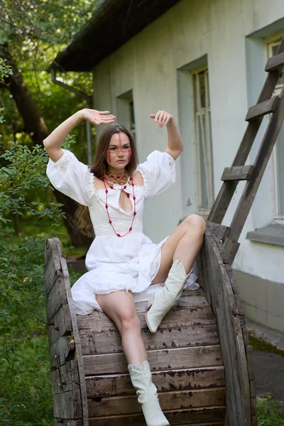 Kırmızı kabile makyajlı ve beyaz elbiseli bir kız, eski ahşap bir tekerleğin üzerinde oturuyor, kollarını eski bir evin arka planına dayayıp poz veriyor ve yaz günü açık havada yemyeşil ağaçlar yeşertiyor. 