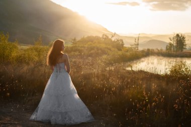 Gün batımında sisli bir manzaraya bakan beyaz elbiseli bir gelinin huzurlu görüntüsü. Doğada hareketsiz duruyor. Huzurlu manzaranın tadını çıkarıyor..