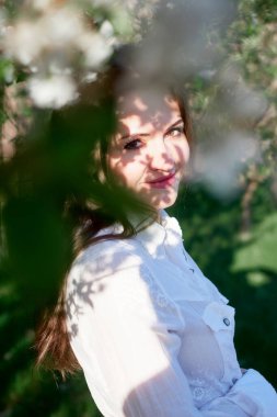 Elma çiçekleriyle çevrili bir bahar bahçesinde güzel bir genç kadın gülümsüyor. Yumuşak güneş ışığı ve gölgeler rüya gibi ve romantik bir atmosfer yaratır..