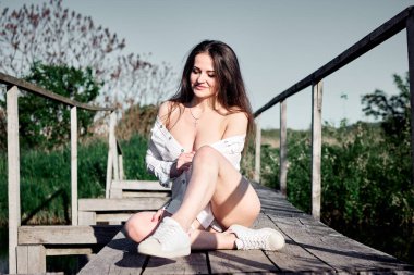 Beyaz gömlekli ve spor ayakkabılı genç, güzel bir esmer tahta bir köprüde bağdaş kurmuş, tatlı tatlı gülümsüyor. Etrafı doğa ile çevrili ve huzurun ve sessizliğin tadını çıkarıyor..