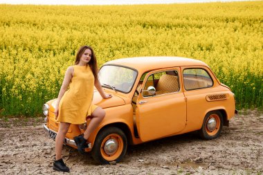 Sarı elbiseli genç bir kadın kolza tohumu tarlasında klasik turuncu bir arabayla poz veriyor ve nostaljik ve renkli bir sahne yaratıyor. Arabanın yanında duruyor..