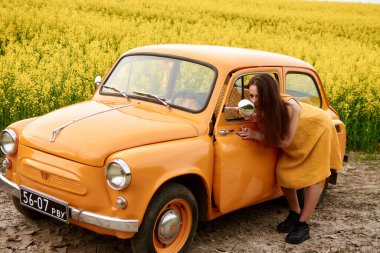 Sarı elbiseli genç bir kadın, eski turuncu bir arabanın yan aynasında kendine hayran, sarı çiçeklerden oluşan canlı bir alana kurulmuş, nostalji ve cazibe hissi uyandırıyor.. 