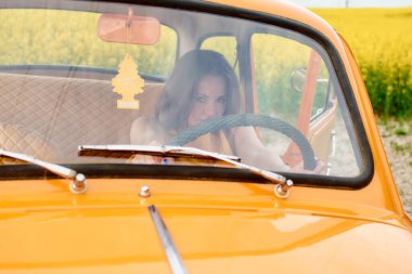 Bir kadın sarı çiçek tarlasında eski turuncu bir araba kullanıyor, cam açık ve silecekler görünür, nostalji ve macera hissi var.. 