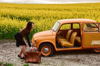 Bulutlu bir günde kolza tohumu tarlasında klasik bir arabanın yanında bavul almak için eğilen bir kadın. Kadın siyah bir elbise giyiyor ve araba turuncu..