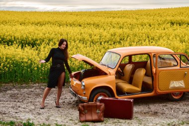 Siyah elbiseli zarif bir kadın kaputu açık turuncu bir arabanın yanında duruyor, bavulları yanında, güzel bir kolza tohumu tarlasında. Geçmişle doğayı harmanlayan bir sahne..