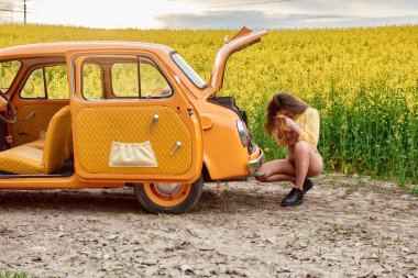 Ayakkabı giyen çıplak bir kadın ve turuncu bir arabanın bagajına çömelmiş sarı bir gömlek, arka planda kolza çiçeği tarlası, güneşli bir günde kır manzarası..