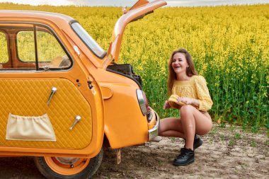 Sarı bluz giyen gülümseyen bir kadın, açık bagajlı turuncu bir retro arabanın yanında çömeliyor. Arka planda sarı çiçekli bir kolza tohumu tarlası var..