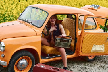 An attractive woman sits in an orange retro car, holding a vintage radio, against a background of a yellow rapeseed field. A concept of summer travel in retro style and vintage traveling.