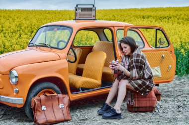 A woman sits with a vintage camera by an orange retro car in a yellow field, a nostalgic roadside scene with a radio on the car's roof and a leather bag nearby. 