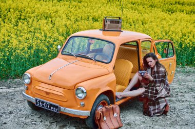 A pretty girl with an old camera in her hands sits near an orange vintage car, on the roof of which there is a radio, surrounded by a field of flowering rapeseed .