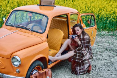 A young woman wrapped in a warm blanket sits in an orange vintage car, holding an old camera. The backdrop features a vibrant yellow rapeseed field, creating a nostalgic and picturesque scene.