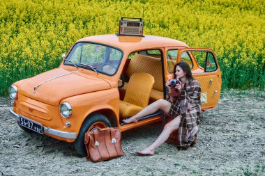 A barefoot woman wrapped in a blanket sits in an orange retro car in a field of yellow flowers with a vintage camera in her hands and a radio on the roof against the backdrop of nature.