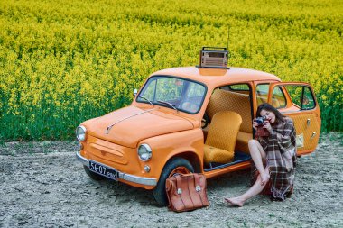 A young woman in a vintage car is taking pictures with a retro camera. She is wrapped in a blanket. The car is parked in front of a yellow flowers field. On the car roof is retro radio.