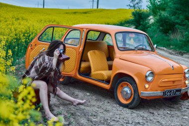 A young woman with an old camera sits on a suitcase next to a vintage car in a field of yellow flowers, capturing the rural charm. A stylish moment of nostalgia and travel.