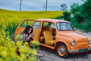 A woman with a vintage camera sits by an orange retro car in a rapeseed field, capturing a rural scene on a sunny day. This picturesque setting evokes nostalgia and a love for photography.