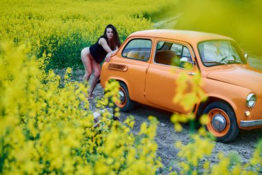 A sensual woman poses near a vintage car in a canola field, surrounded by yellow flowers, creating a captivating scene of beauty and classic style in a rural landscape.