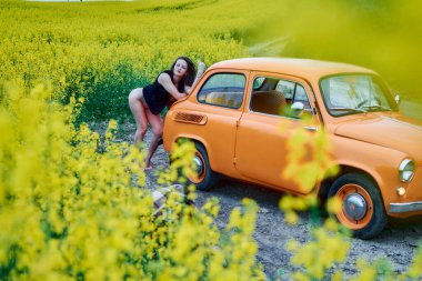 A beautiful brunette woman is posing seductively next to a vintage orange car in a vibrant yellow canola field. It's a sunny day, and the scene exudes a sense of freedom and nostalgia.