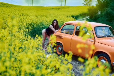 A beautiful brunette poses near an orange car in a vibrant yellow rapeseed field, capturing a moment of natural beauty and elegance on a sunny day.