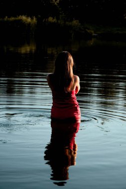 A woman stands waist-deep in calm water at sunset, her back to the camera and her reflection visible on the lake, creating a serene and solitary moment.