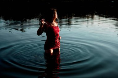 A stunning image of a young woman in a red dress standing in a serene lake at sunset, creating a dreamlike atmosphere and radiating peace and tranquility. The ripples in the water add a dynamic touch.