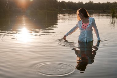 A young woman in a pink top and white shirt stands in the lake at sunset, gently touching the water's surface. The sun sets over the horizon, reflecting on the water. 