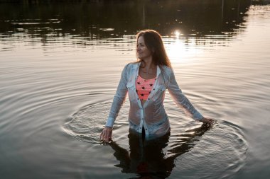 A young woman stands in a lake at sunset, wearing a wet shirt and swimsuit. She smiles as she touches the water, creating ripples and enjoying the tranquil scene. 