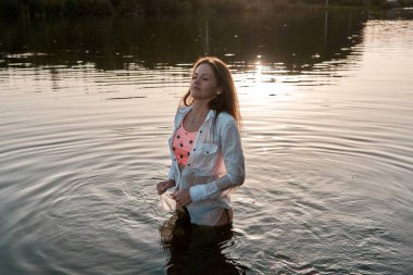 A serene young woman stands peacefully in a lake at sunset, wearing a white shirt and swimwear, feeling the water and enjoying the tranquil moment in the golden light. 