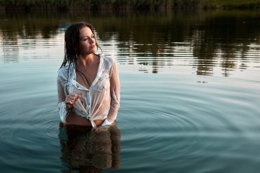A woman stands waist-deep in a lake, wearing a wet white shirt. The scene is bathed in soft light, highlighting her sensual and serene expression amidst the tranquil natural setting.
