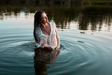 A woman stands waist-deep in a lake, wearing a wet white shirt. The sunset casts a warm glow as she looks away peacefully. The image conveys serenity, natural beauty and peacefulness.