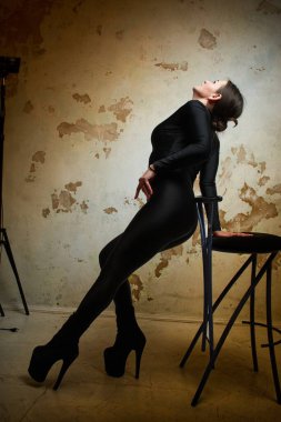 An elegant woman in a black catsuit poses gracefully on a chair, creating a striking contrast against a textured grunge wall, highlighted by dramatic lighting for a chic, artistic effect.