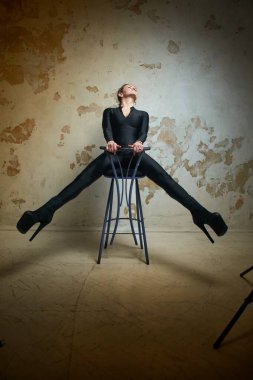 A striking studio portrait of a woman in a sleek black bodysuit, seated on a stool with her legs spread, creating a powerful and confident pose against a textured background. 