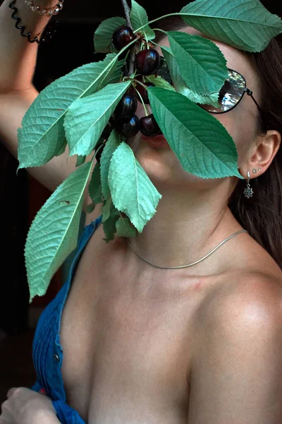 A beautiful woman with bare shoulders poses with a branch of ripe cherries in front of her face, the leaves partially obscuring her features, creating an intriguing and sensual atmosphere .