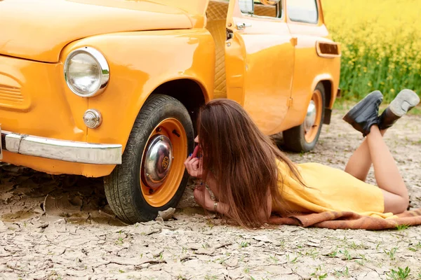 Turuncu elbiseli, uzun saçlı çekici bir kız eski turuncu bir arabanın yanında yatıyor ve yazın sarı çiçeklerle dolu bir tarlada arabanın direksiyonunu yalıyor..