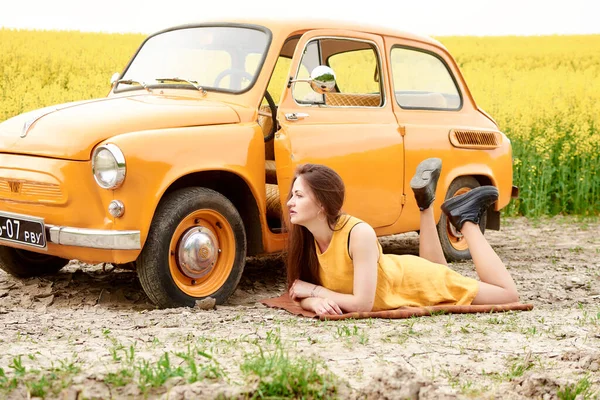 Sarı elbiseli genç bir kadın, arka planda sarı çiçekli kırsal bir alanda turuncu bir arabanın yanında poz veriyor, canlı ve nostaljik bir sahne yaratıyor..