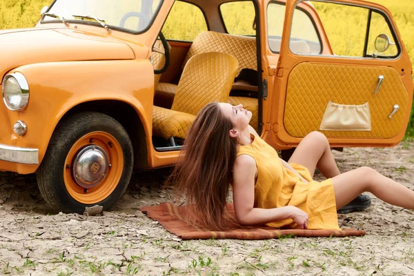 Sarı elbiseli bir kadın, sarı çiçeklerle dolu bir tarlada turuncu bir arabanın yanında battaniyede dinleniyor. Poz veriyor ve arka planda açık bir araba kapısıyla yukarı bakıyor..