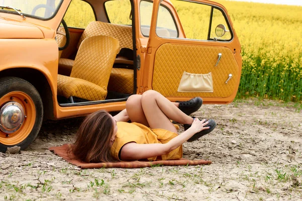 Sarı elbiseli genç bir kadın turuncu bir arabanın yanındaki halıya uzanmış, kapısı açık, arka planda sarı kolza çiçeği tarlası, yaz tatili..