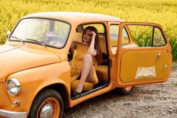 Güzel bir kadın, arka planda kolza tohumu tarlası olan klasik turuncu bir arabada poz veriyor. Yaz zarafeti ve cazibesini yakalıyor.. 