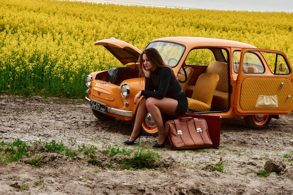 Siyah elbiseli bir kadın, sarı çiçekli bir tarlada kapüşonlu eski bir arabanın yanında bavulunun üzerinde oturuyor. Sıkıntılı görünüyor, yardım bekliyor ya da kırsalda tamirci bekliyor..