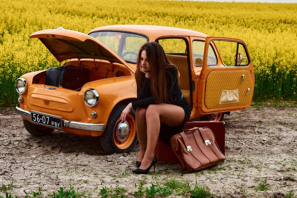 Siyah elbiseli genç bir kadın kaputu açık turuncu bir arabanın yanında bir valizin üzerinde oturuyor. Araba yolda bozuldu ve o kolza tohumu tarlasında yardım bekliyor..