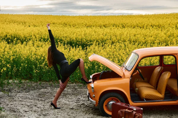 Siyah elbiseli şık bir kadın sarı bir çiçek tarlasında eski bir turuncu arabanın yanında poz veriyor. Sahne, modayla otomotiv estetiğini doğal ortamda birleştiriyor.. 