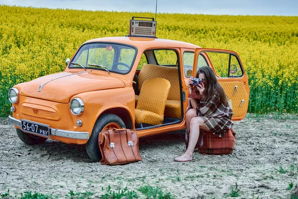 A vintage orange car is parked in a field of yellow flowers, with a young woman sitting on a suitcase and taking a photo with an old camera, creating a nostalgic and charming scene.