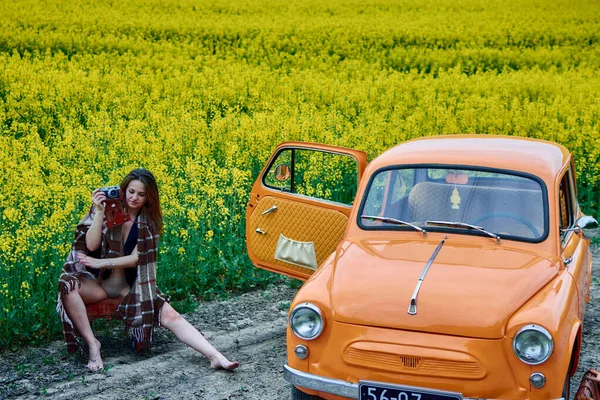 A woman in a plaid sits next to an orange vintage car in a rapeseed field, holding a film camera, capturing a nostalgic and timeless moment in the countryside. 