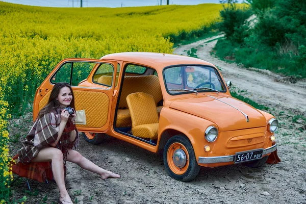 A young woman sits with a camera next to an orange vintage car on a dirt road, with a field of yellow flowers in the background, capturing a moment of rural beauty and timeless charm.