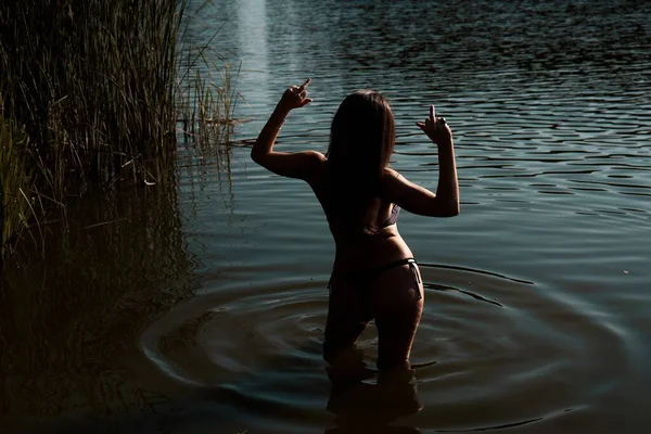 A back view of a young woman in a swimsuit showing middle finger gesture in the lake surrounded by reeds during summer vacation. Freedom and defiance concept. 