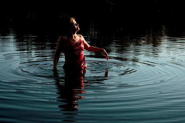 A young woman in a red dress stands in a lake, her face turned upwards to the sky, with her right hand gently touching the surface of the water, creating a serene and contemplative mood.