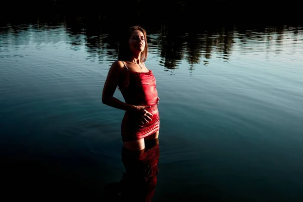 A woman in a red dress standing in water, lake water in the evening sun. Seductive pose, light plays, reflection of the dress in the water. A beautiful and sensual scene. 