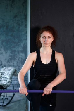 The woman in sportswear is doing a stretching exercise with a purple bar in her hands in the studio against a dark wall, looking straight to the camera with a serious expression on her face.